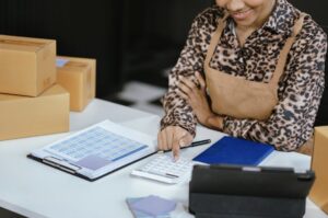 A business owner using a calculator and tablet to manage wholesale bookkeeping for a distribution company with shipping boxes in the background.