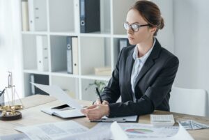 An attorney in a suit and glasses, analyzing financial documents and performing trust account reconciliation at a desk with a calculator, money, and a brass scale of justice.